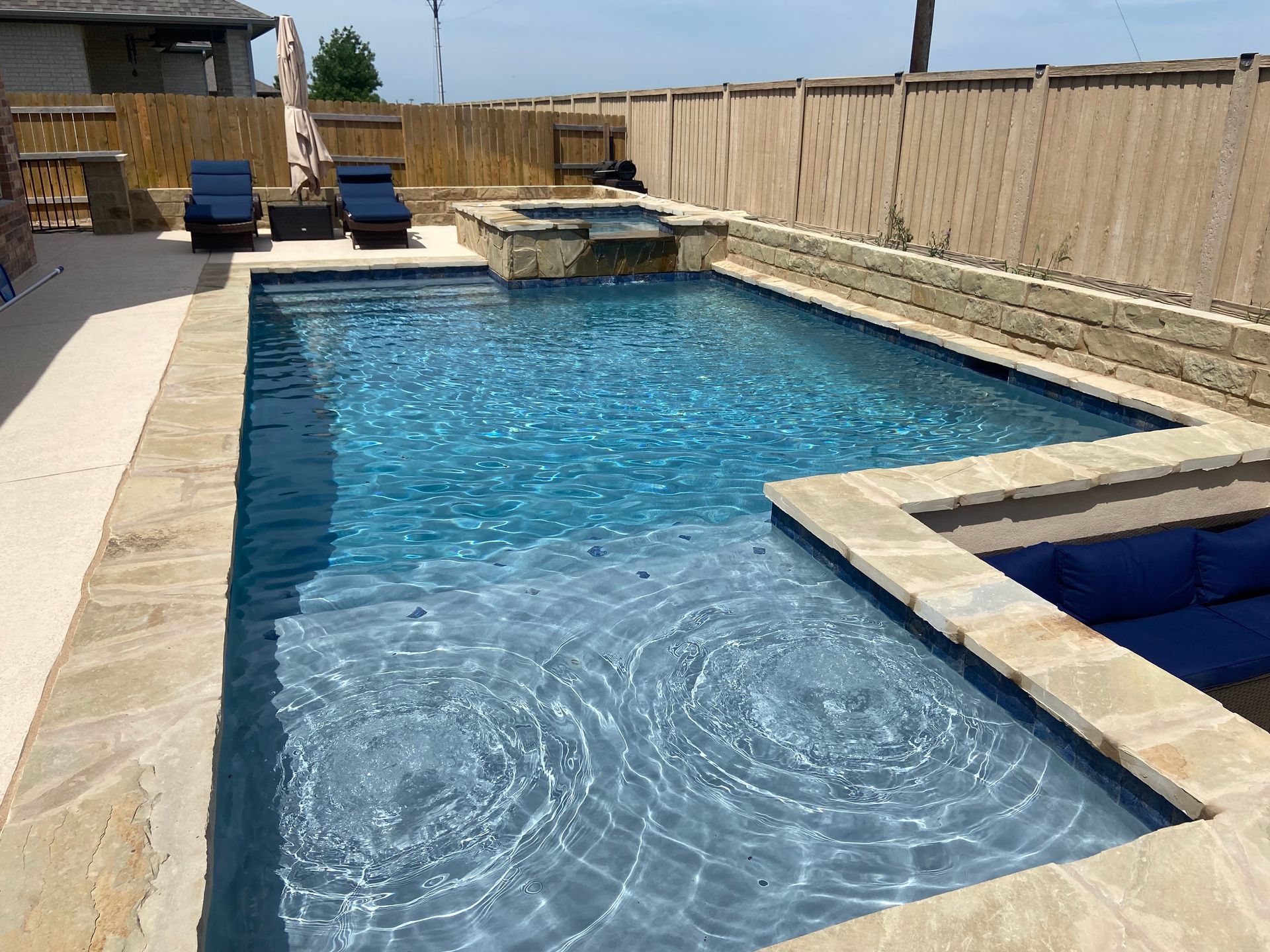 Rectangular pool with stone border, blue water, hot tub, lounge chairs, and wooden fence.