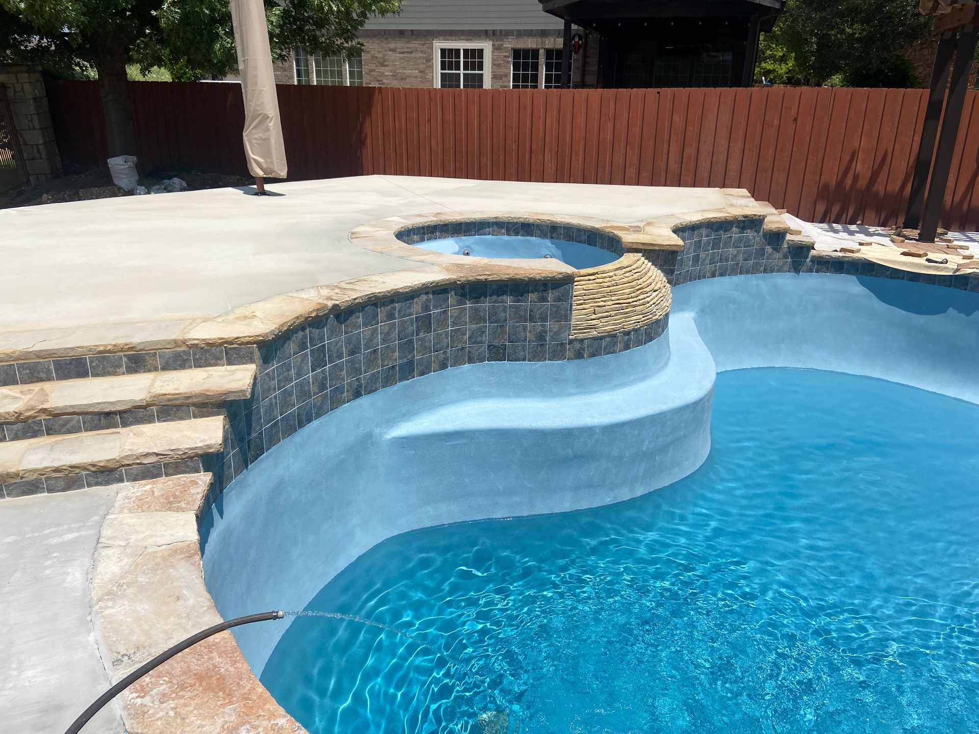 Pool with built-in hot tub, steps, and blue water, surrounded by stone and concrete, with a brown fence in the background.