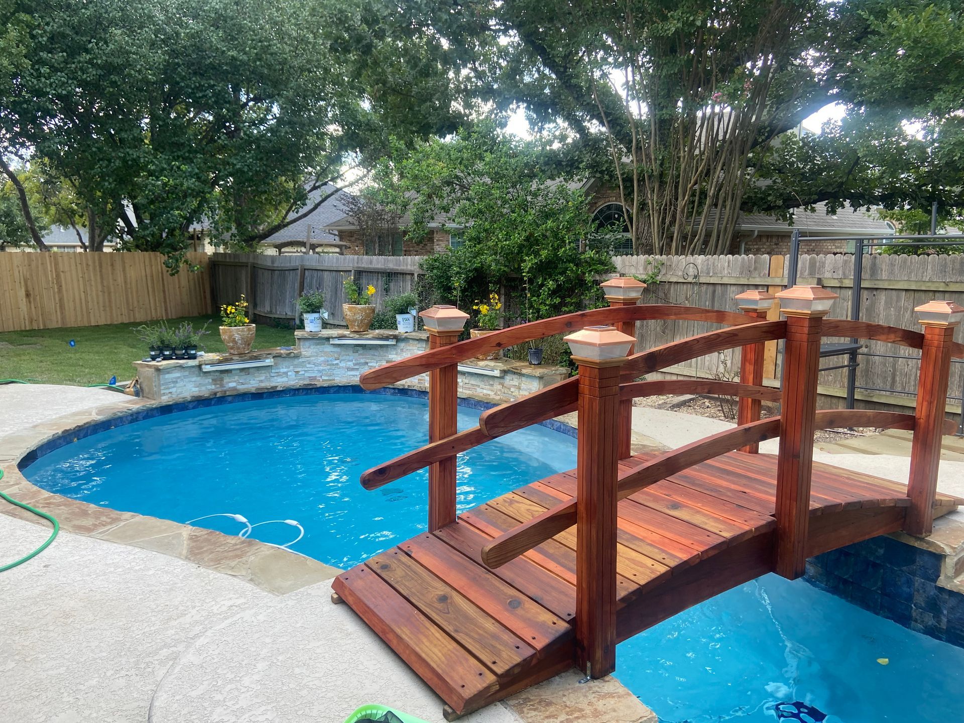 A backyard pool with a wooden bridge, surrounded by a stone deck, green grass, and trees.