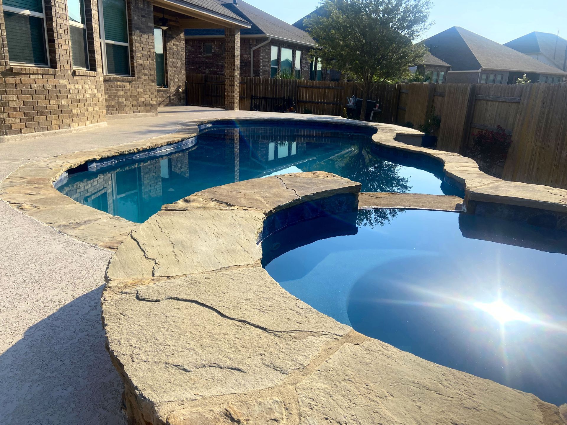 Swimming pool with stone coping and a light-colored deck.  A blue sky and houses are in the background.