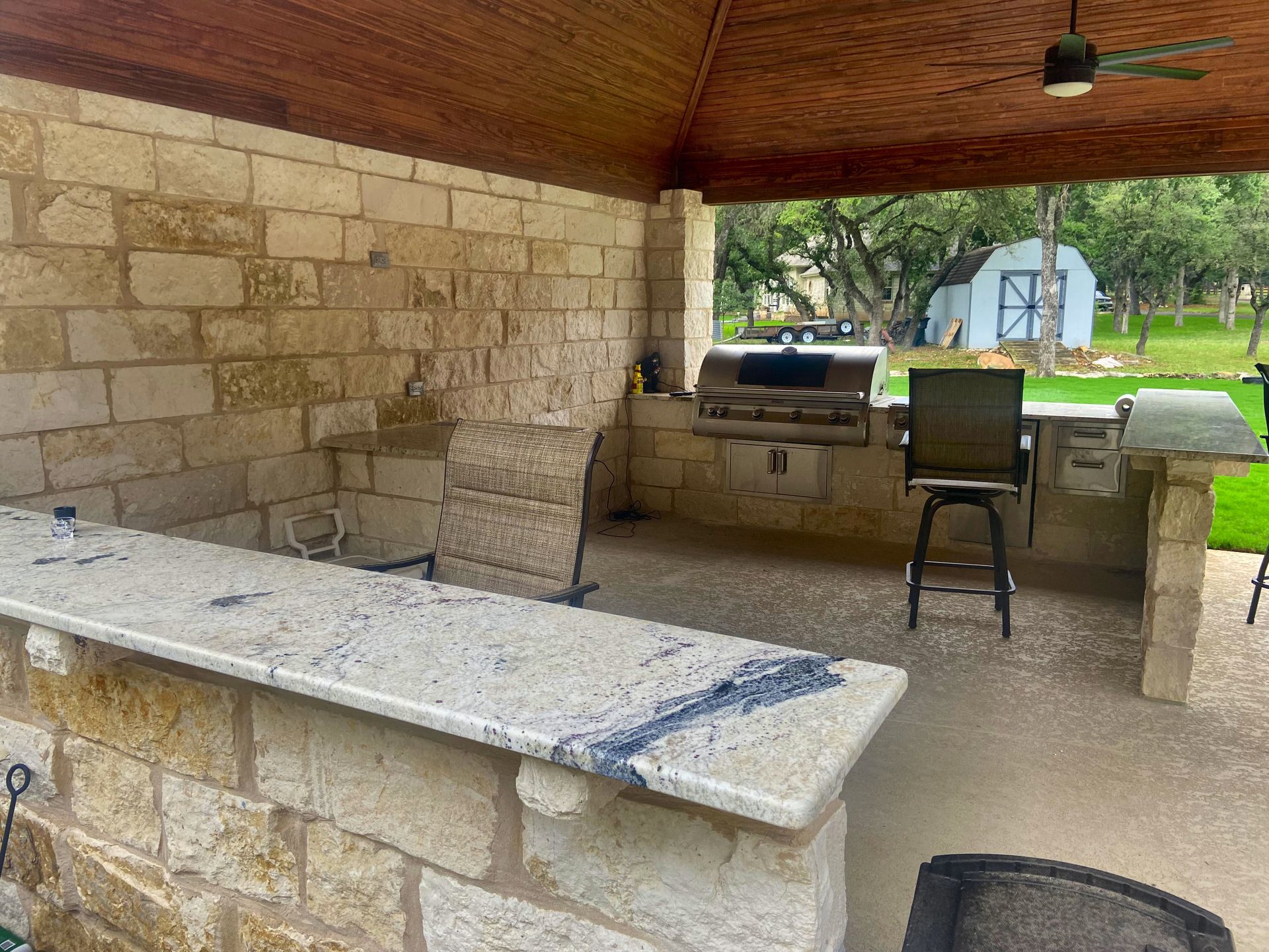Outdoor kitchen with stone walls, granite countertops, grill, and bar stools; shed in background.