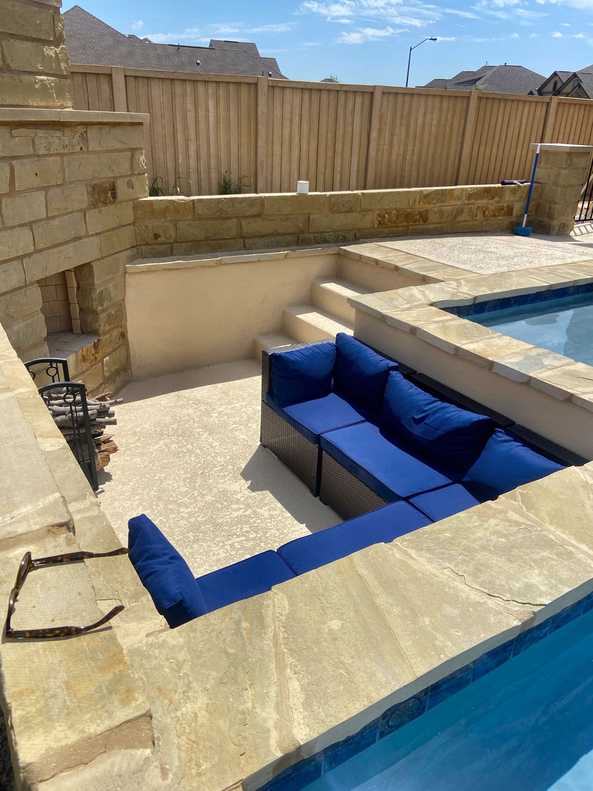 L-shaped blue sofa near a pool with stone steps and patio. Sunny outdoor setting with wooden fence.