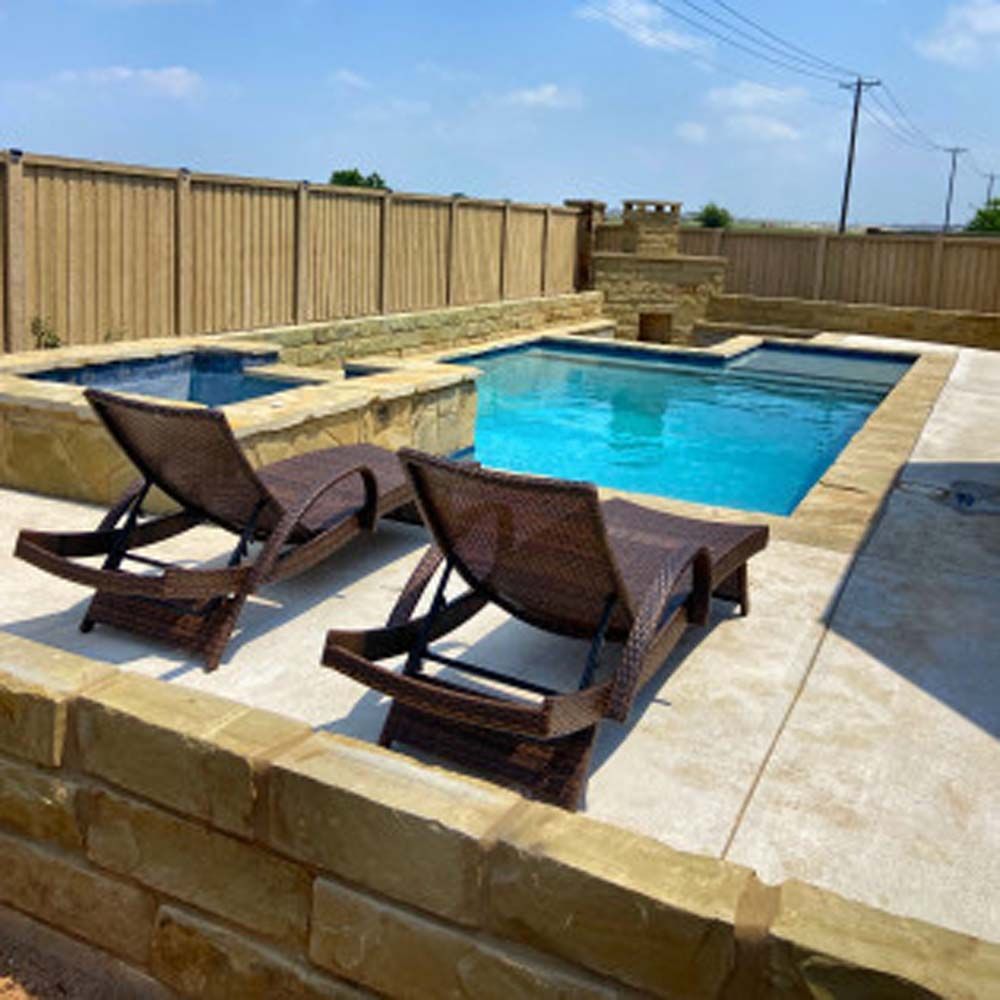 Two brown lounge chairs sit beside a blue pool and jacuzzi, surrounded by a stone wall and wooden fence.