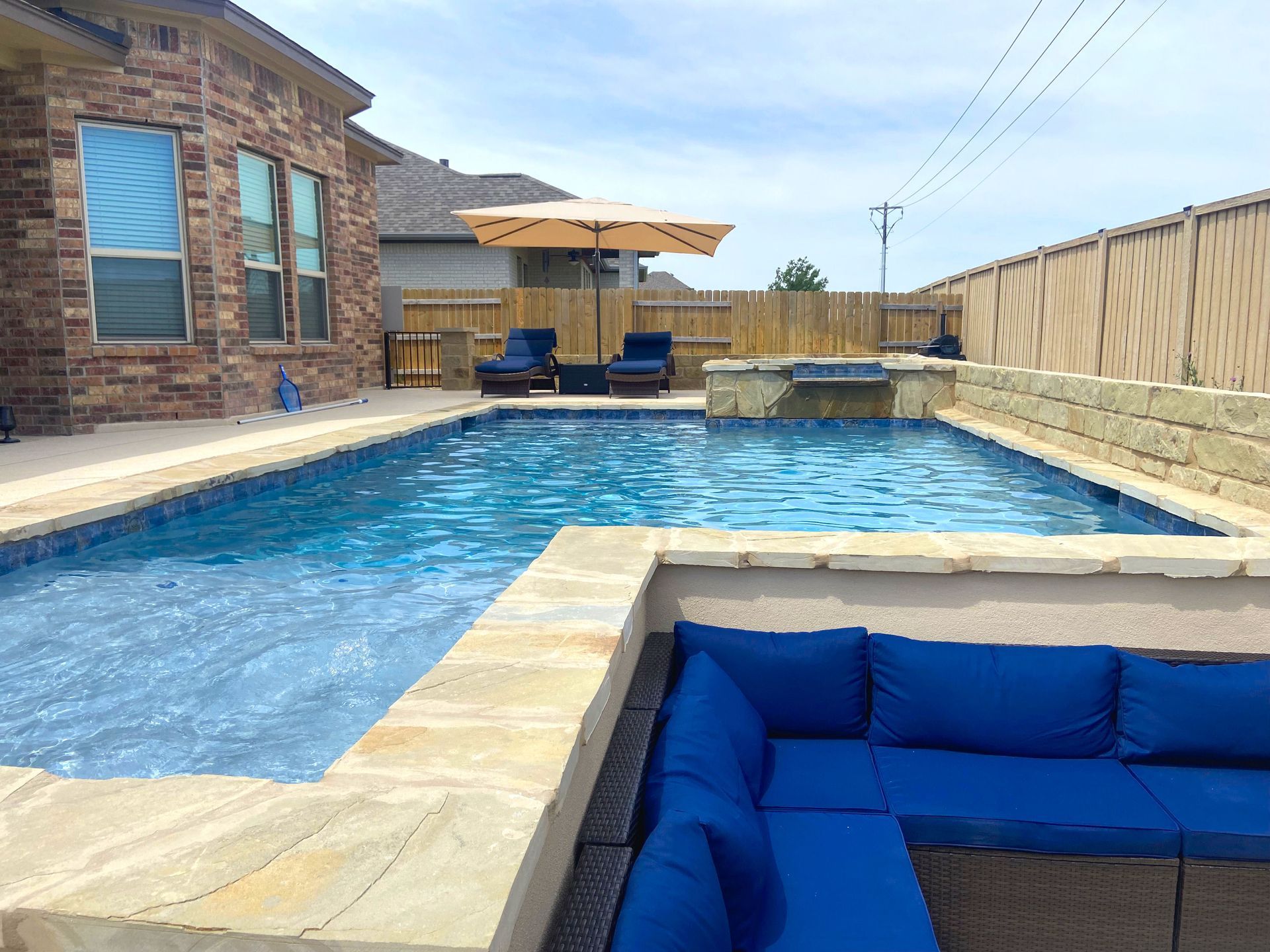Backyard pool with blue cushions, umbrella, lounge chairs, and a brick house.