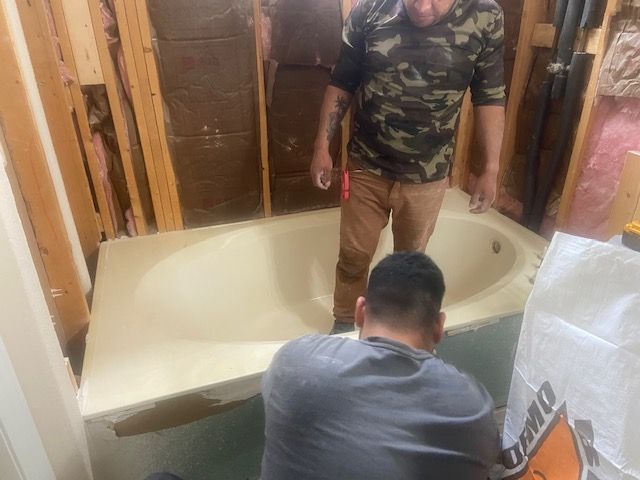 Two men installing a beige bathtub in a partially constructed bathroom; wooden studs visible.