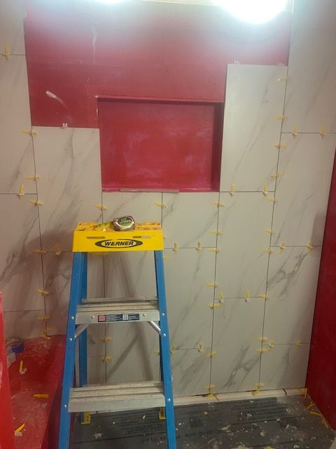 Tile installation in progress; cream tiles with yellow spacers on a red wall, step ladder in foreground.