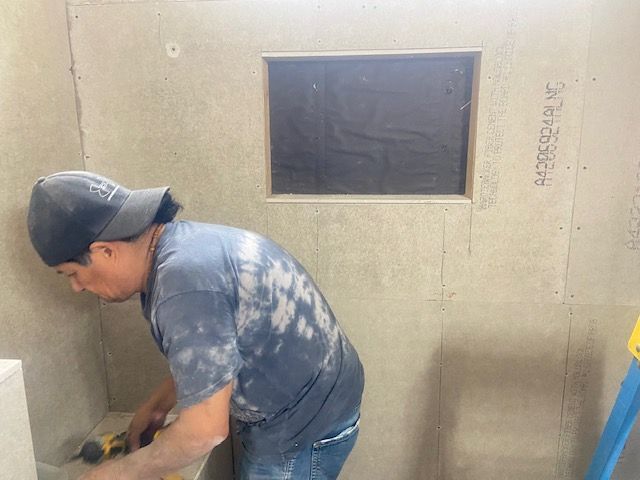Man in a hat drills into shower wall. Gray walls, square window, unfinished interior.