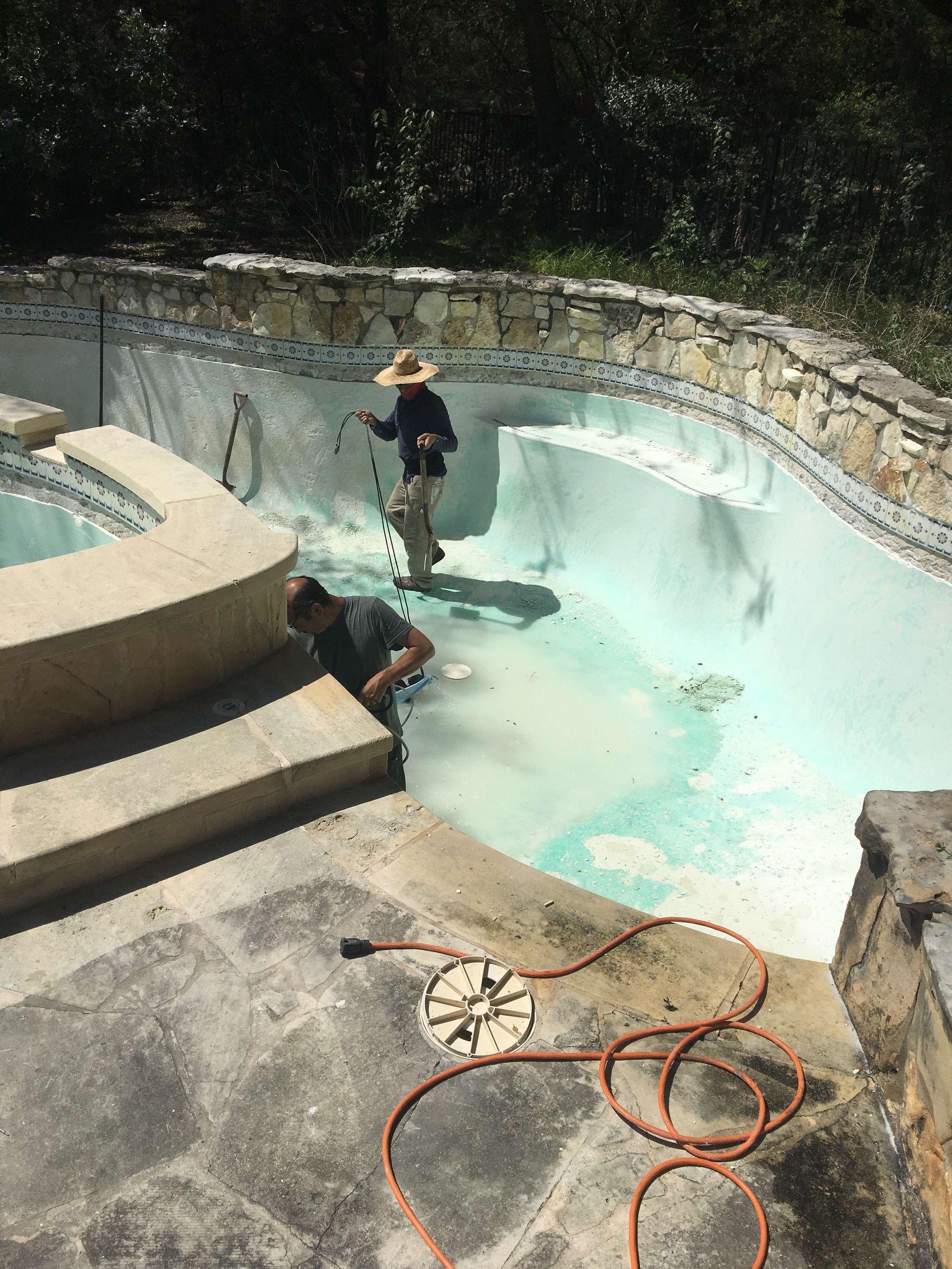 Two people cleaning a drained pool. One uses a hose, the other looks at the water. Outdoor setting with stone accents.