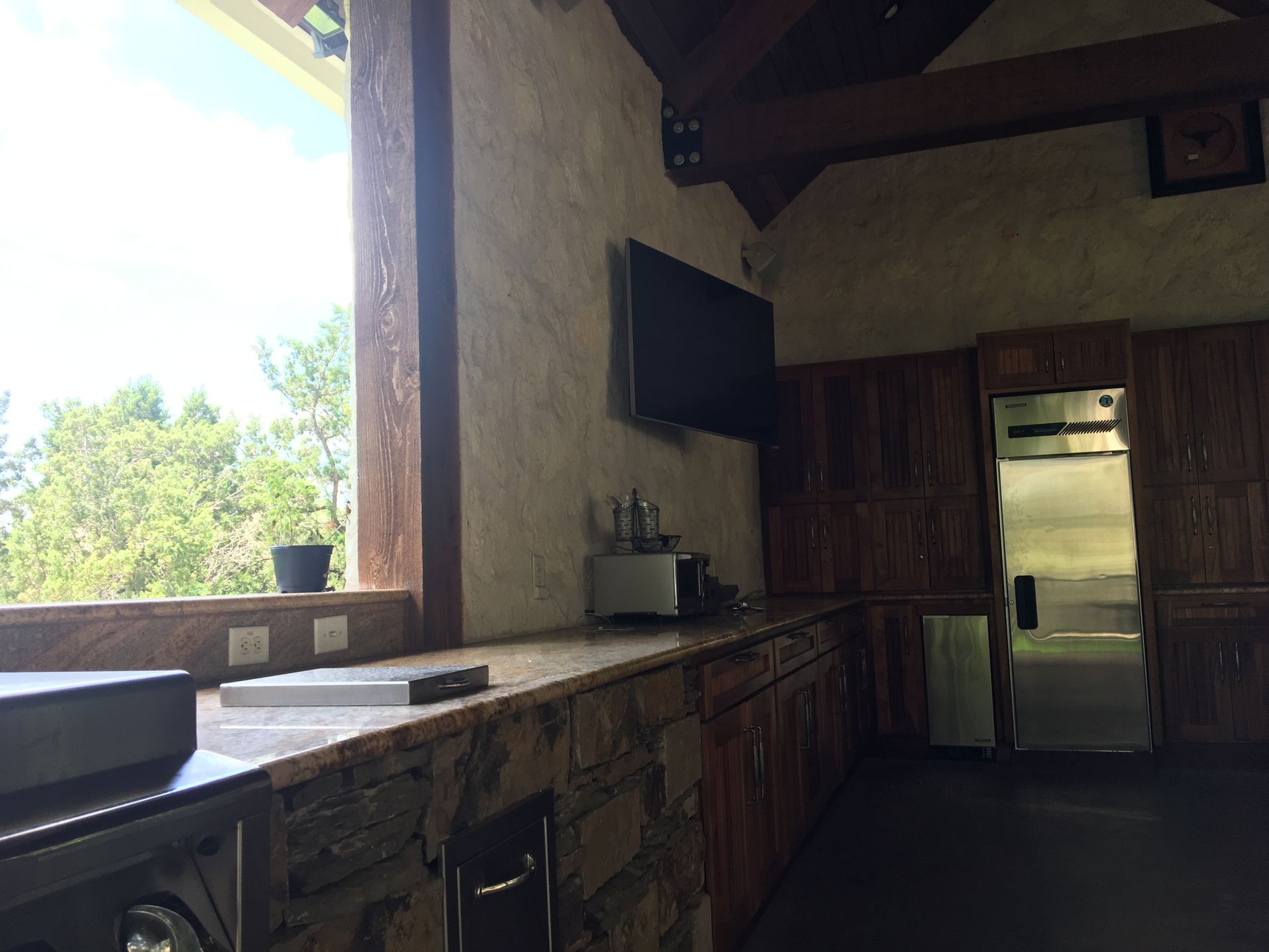 Outdoor kitchen with a window, countertop, cabinets, a TV, and a refrigerator.
