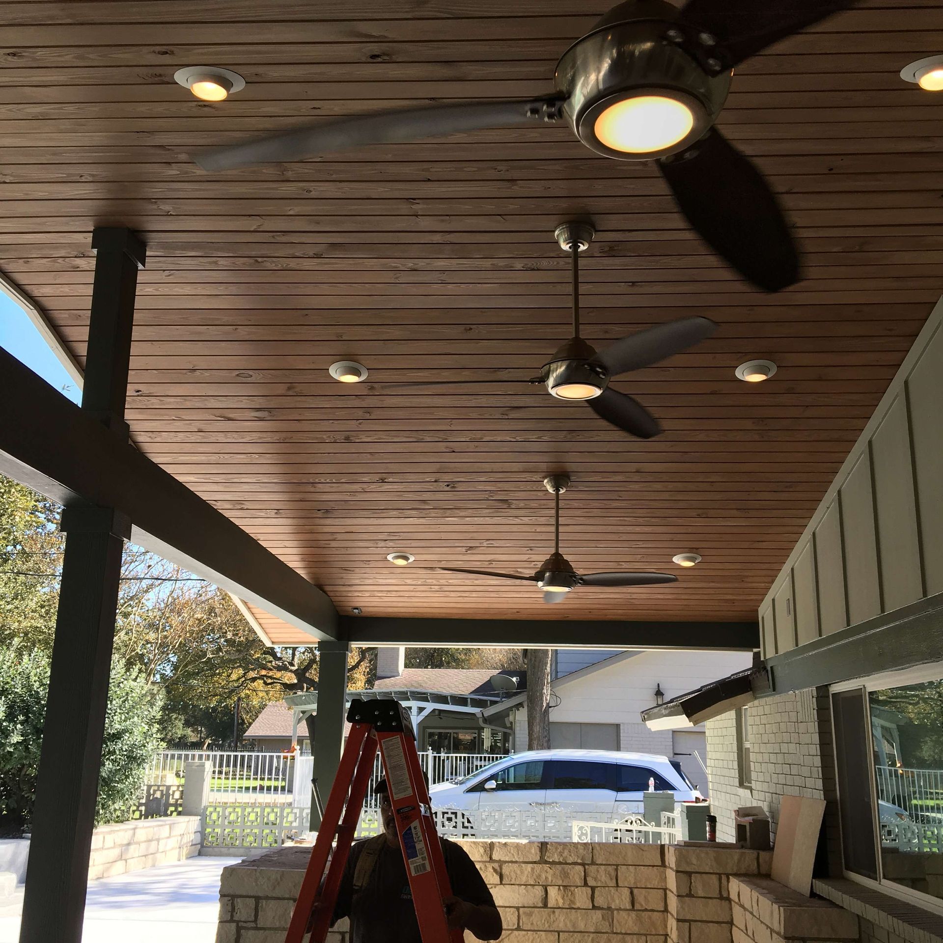 Covered patio with ceiling fans, recessed lights, and a ladder; person on the ladder.