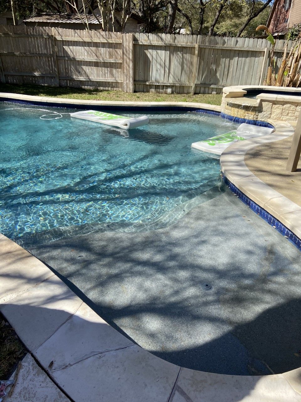 Pool with blue water, steps, and light-colored stone border against a wooden fence.