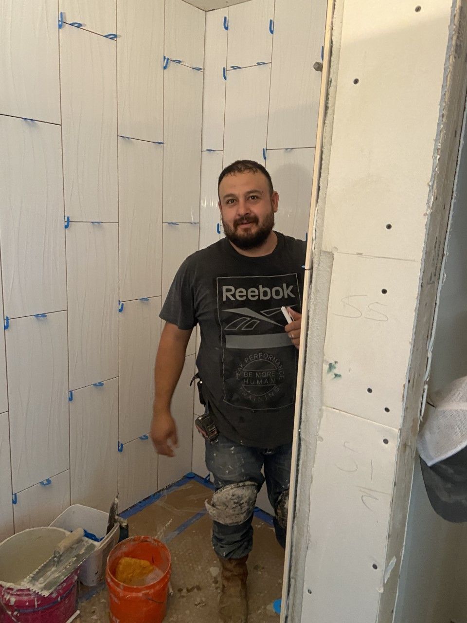 Man in a tiled shower, wearing a Reebok shirt and knee pads. He is holding something and smiling.