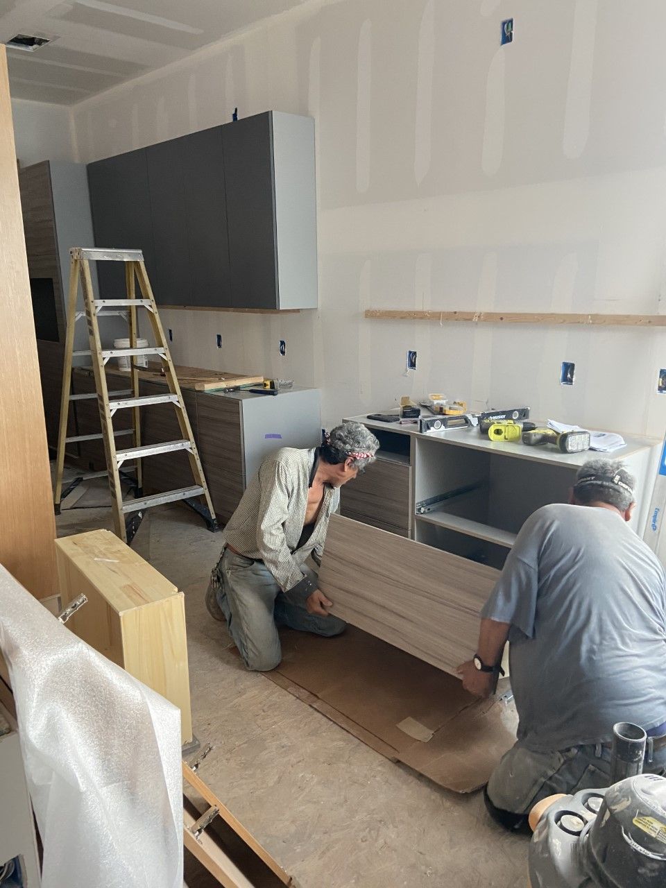 Two workers installing a kitchen cabinet, gray and beige, in a room under construction.
