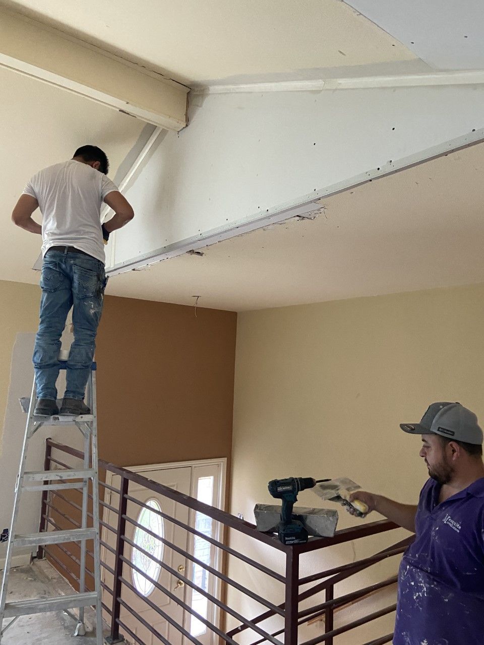 Two men working on a ceiling, one on a ladder, the other holding tools on a railing.