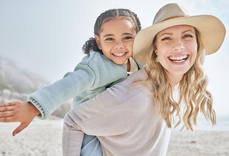 A woman is carrying a little girl on her back on the beach.