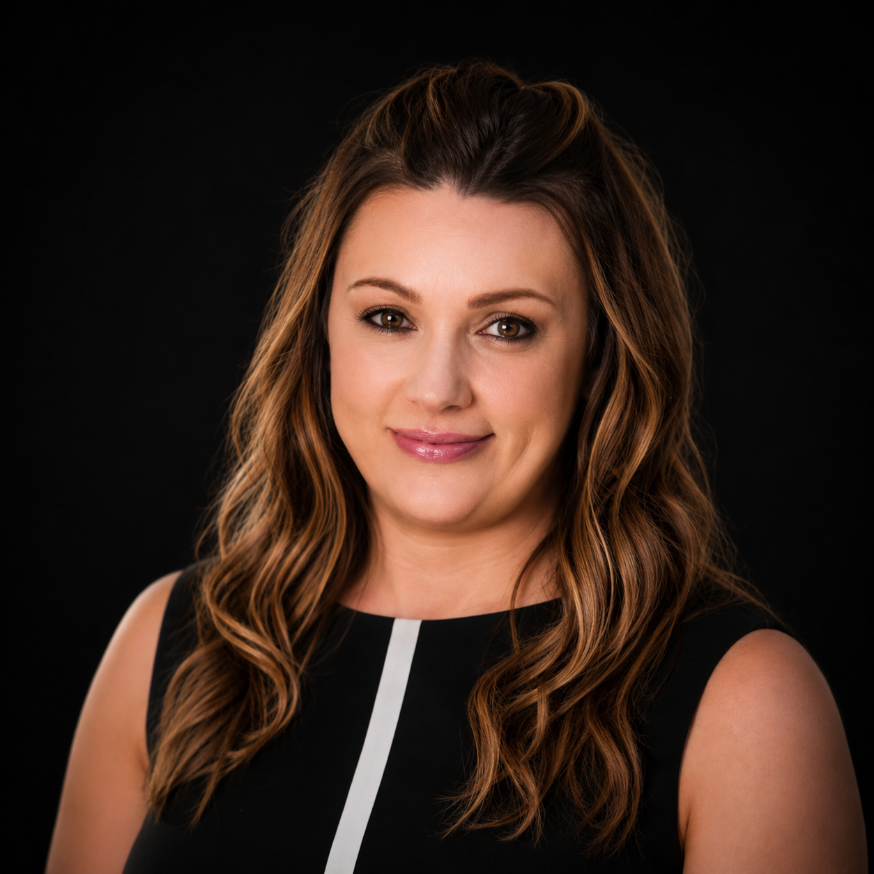 Studio headshot of a smiling woman with long wavy brown hair in a black top on a dark background