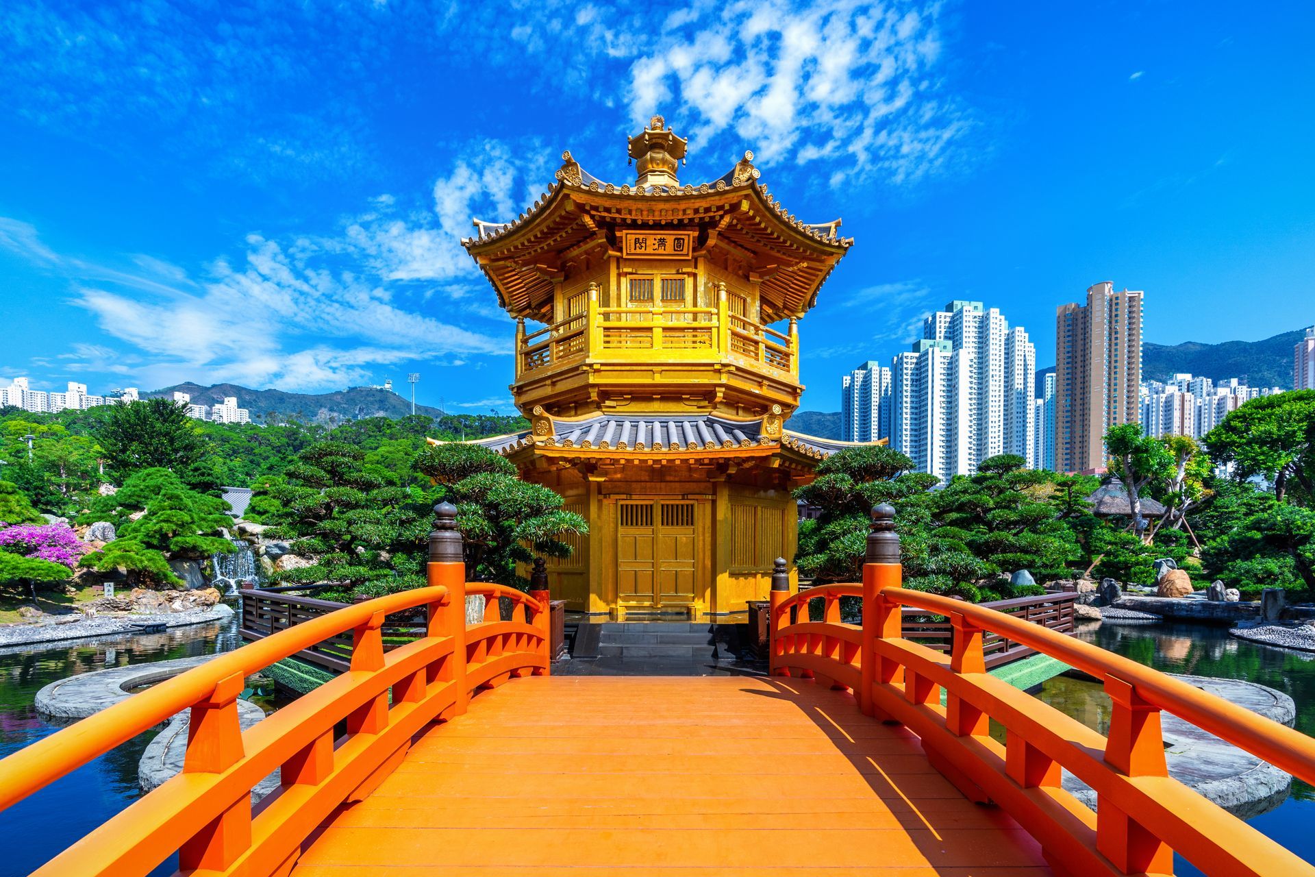 Golden pavilion with orange bridge over water in a park, mountains and skyscrapers in the background, blue sky.