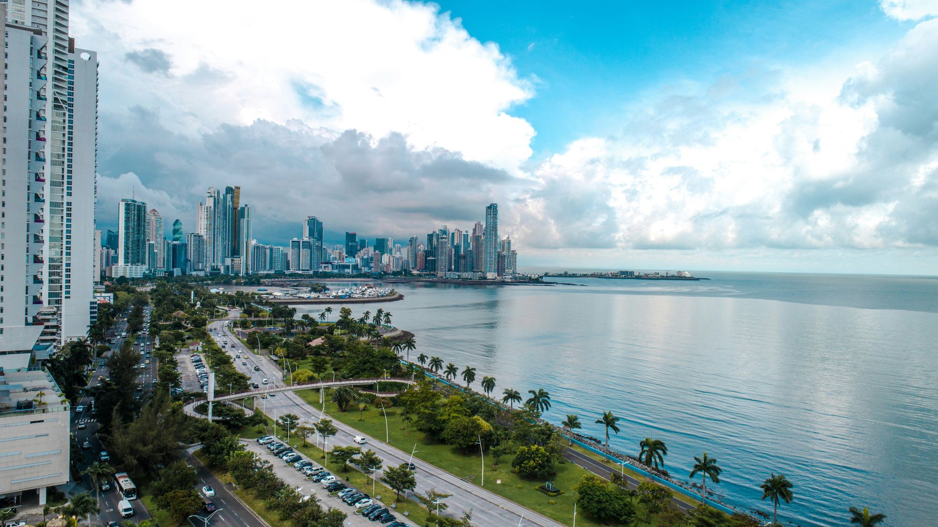 City skyline by ocean under cloudy sky. Tall buildings line waterfront road.