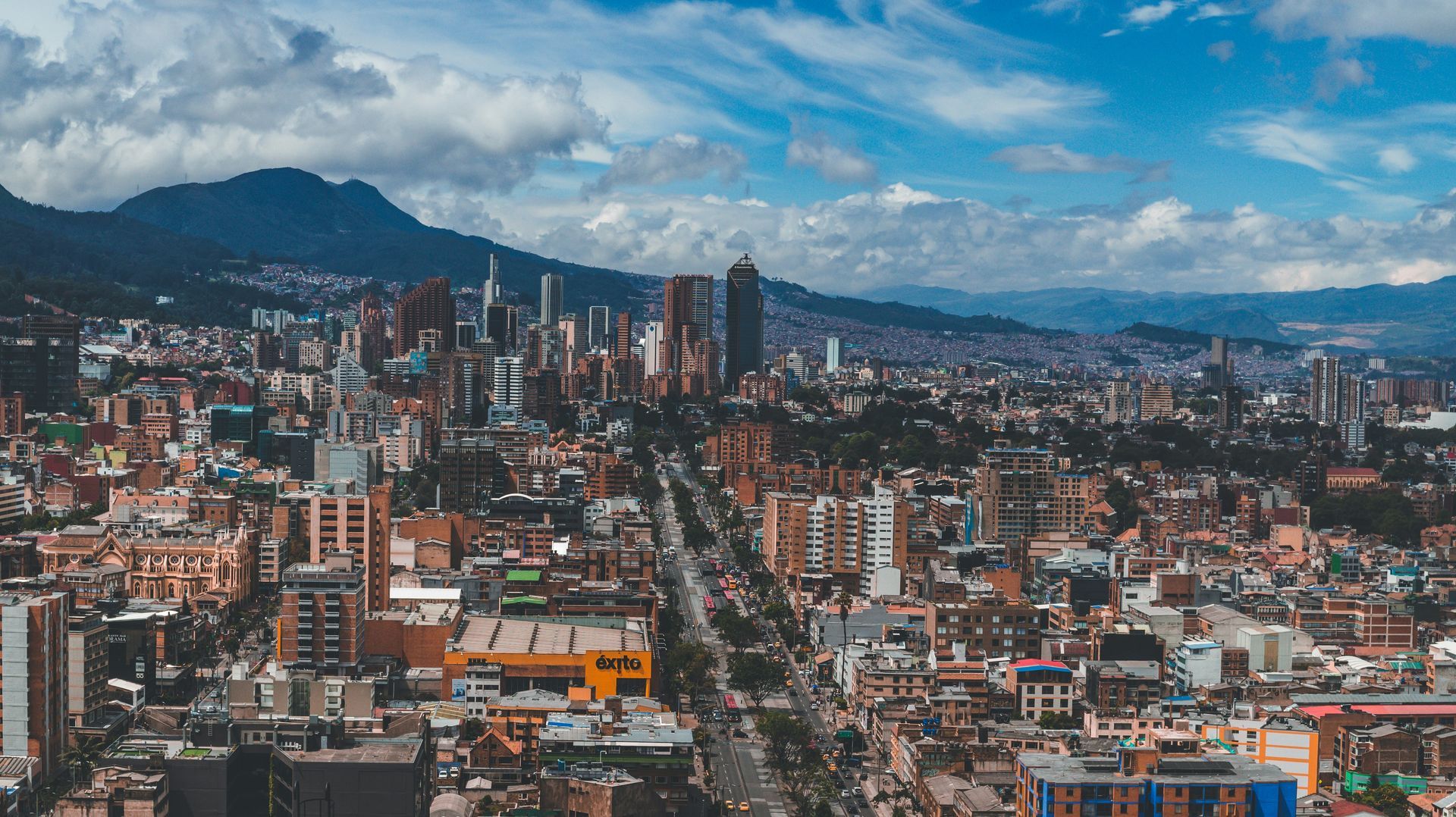 Aerial view of a sprawling cityscape with tall buildings, streets, and mountains in the background under a blue sky.