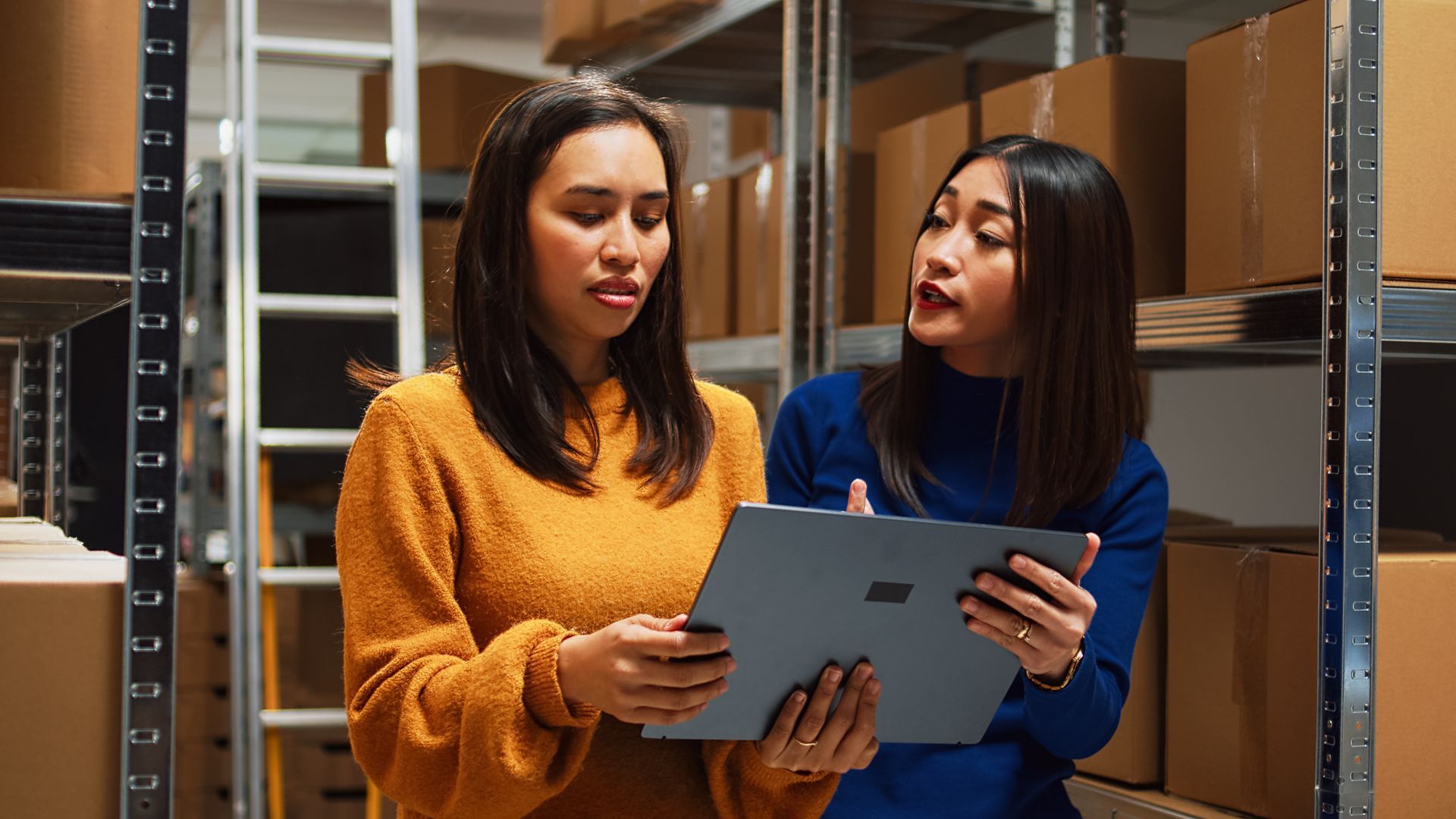 Two women in a warehouse looking at a tablet. One points, the other looks on. Shelves of boxes surround them.