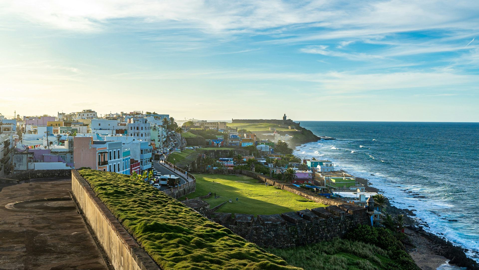Coastal town with buildings, green grass, and ocean under a blue sky.