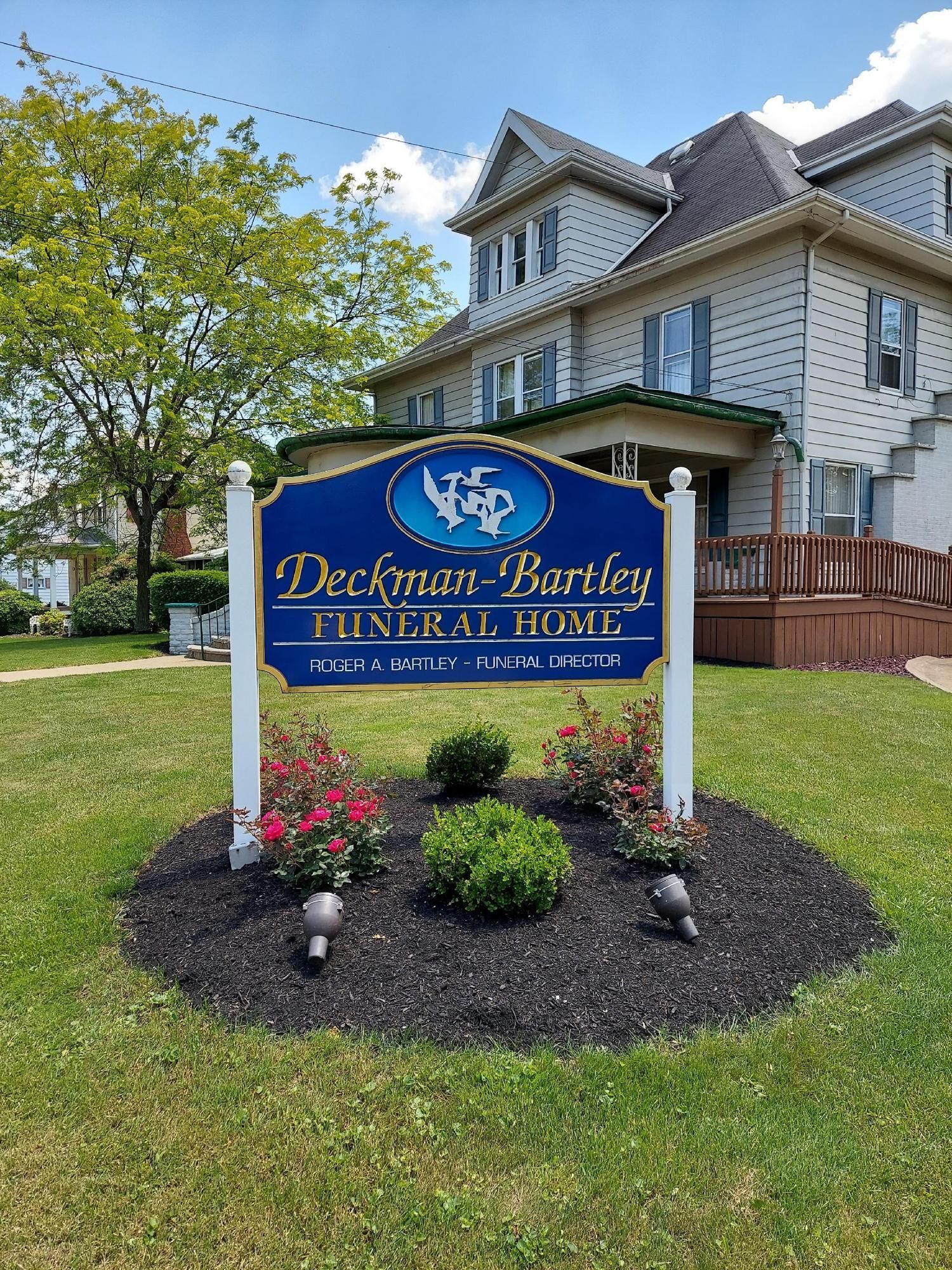Sign for Deckman-Bartley Funeral Home. Blue sign with gold lettering and decorative flowers in front of a large house.