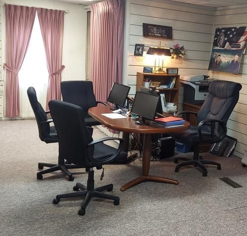 An office with a round table, five black chairs, computers, and a printer. Pink curtains and an American flag photo.