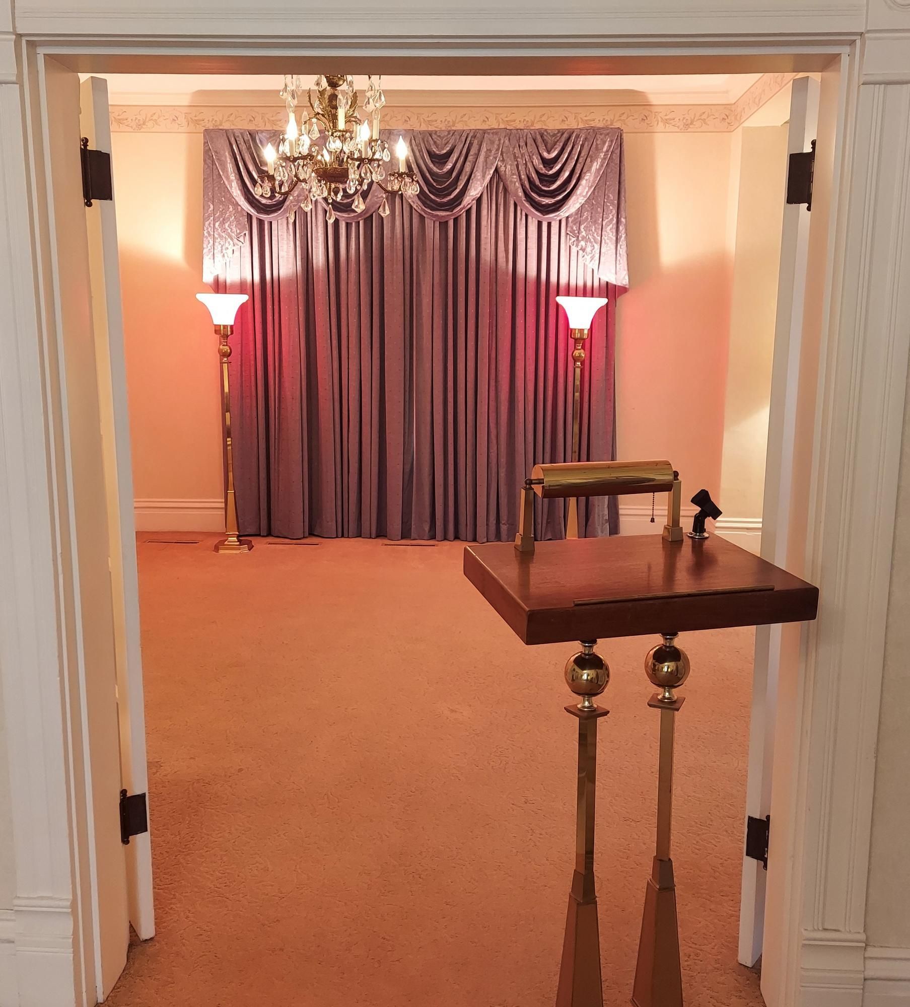 Interior of a room with a curtained backdrop, chandelier, and a podium. Warm lighting and a plush carpet.