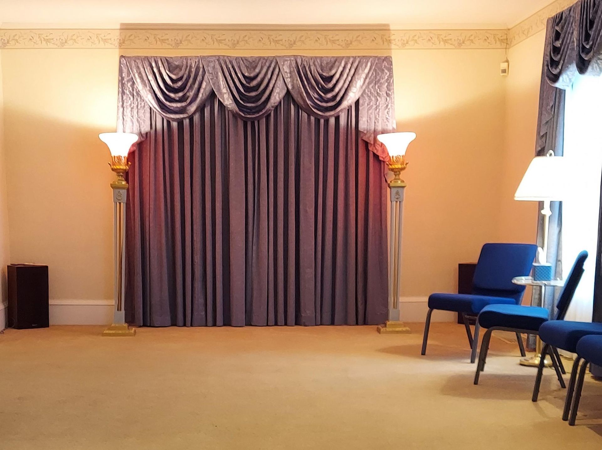 Empty funeral home chapel with mauve curtains and gold-trimmed lamps. Blue chairs are near a window.