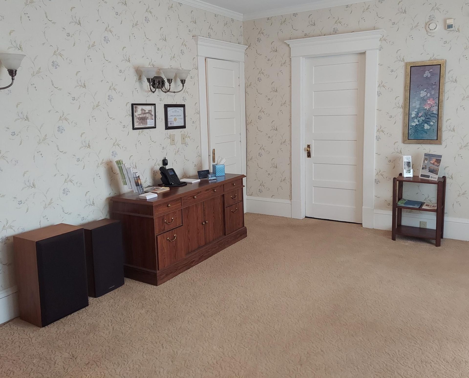 Room with brown furniture, speakers, and floral wallpaper. A desk, two doors, and a small table are present.