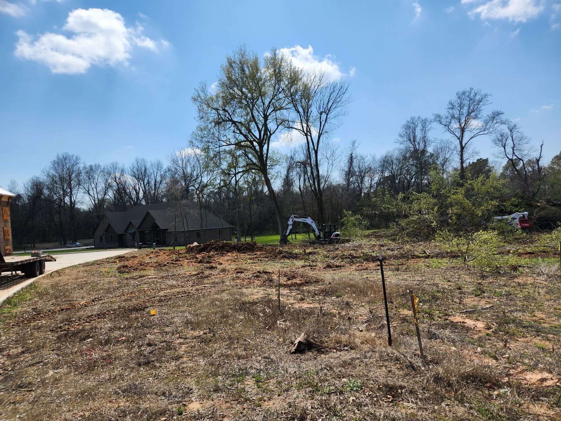 Cleared lot with dirt and small trees, house in the background, excavator in center.