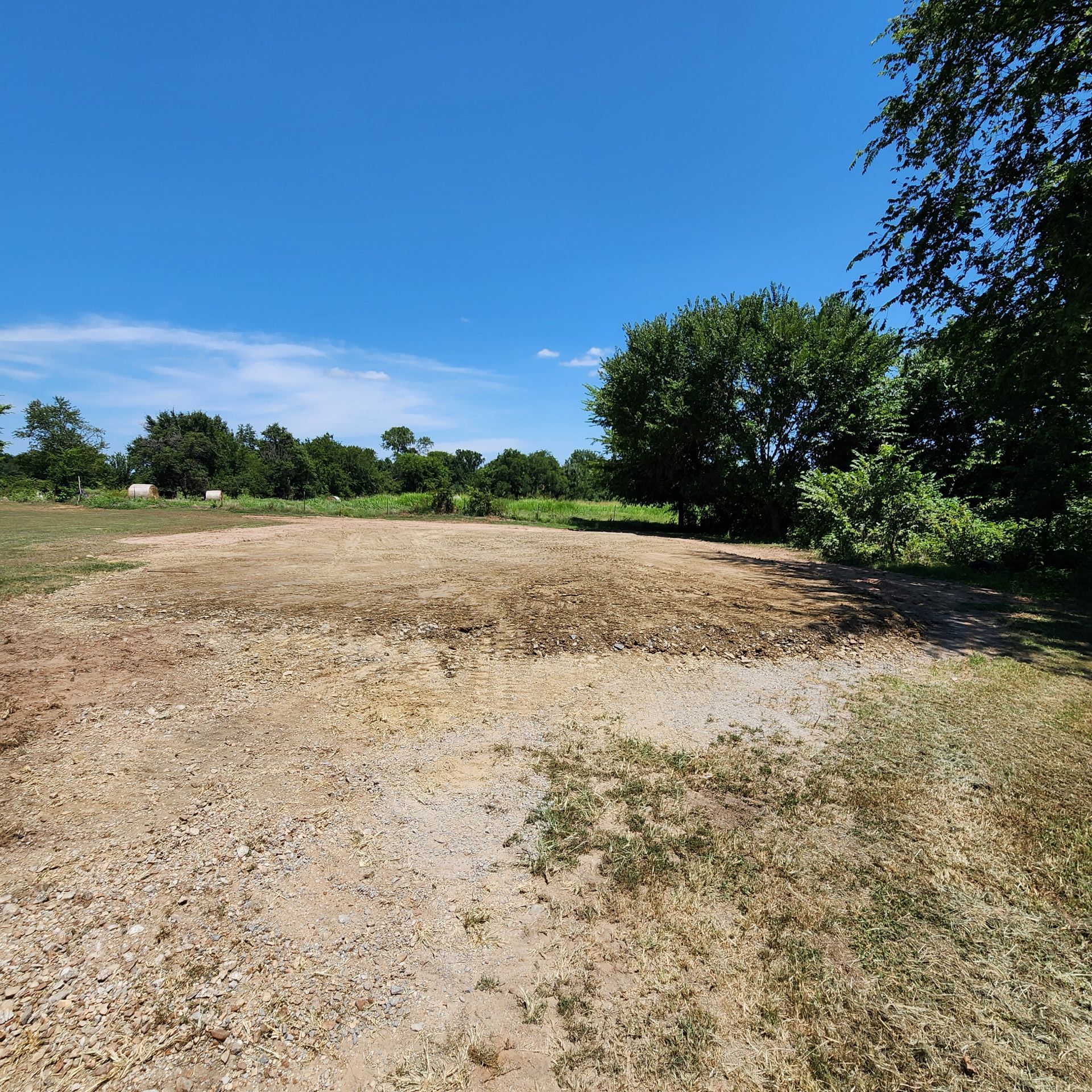Cleared dirt area with gravel and sparse vegetation under a blue sky, trees in the background.