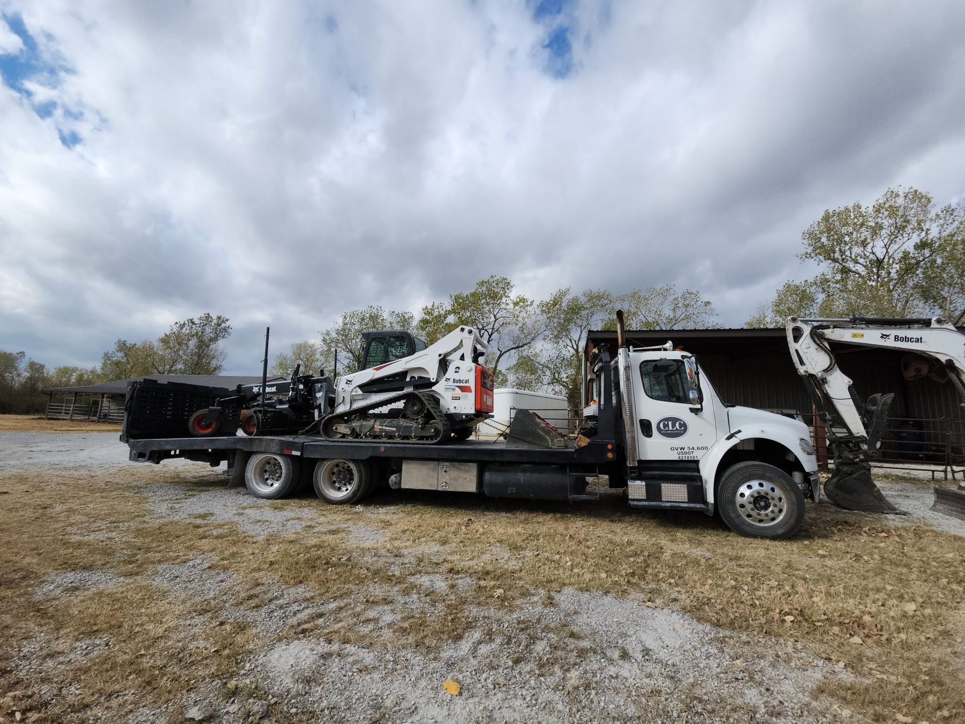 White truck with a flatbed carrying two Bobcat construction vehicles. Overcast sky.