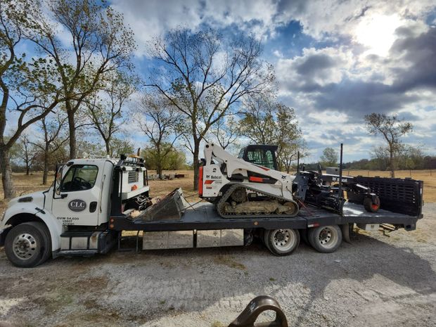 Truck carrying a Bobcat track loader and construction materials on a flatbed trailer outdoors.