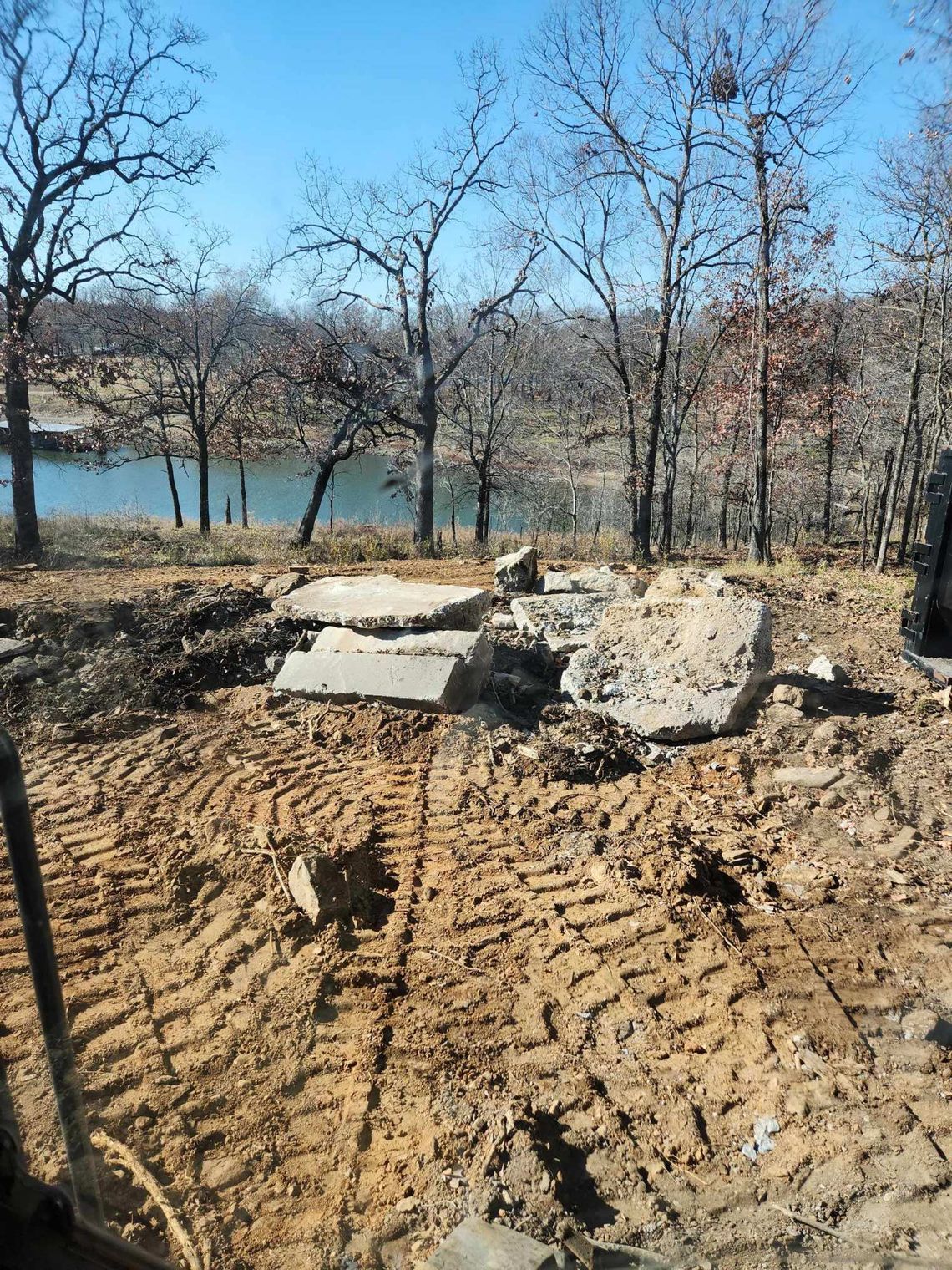 Demolished concrete slabs on dirt, tires tracks, bare trees, and a lake in the background.
