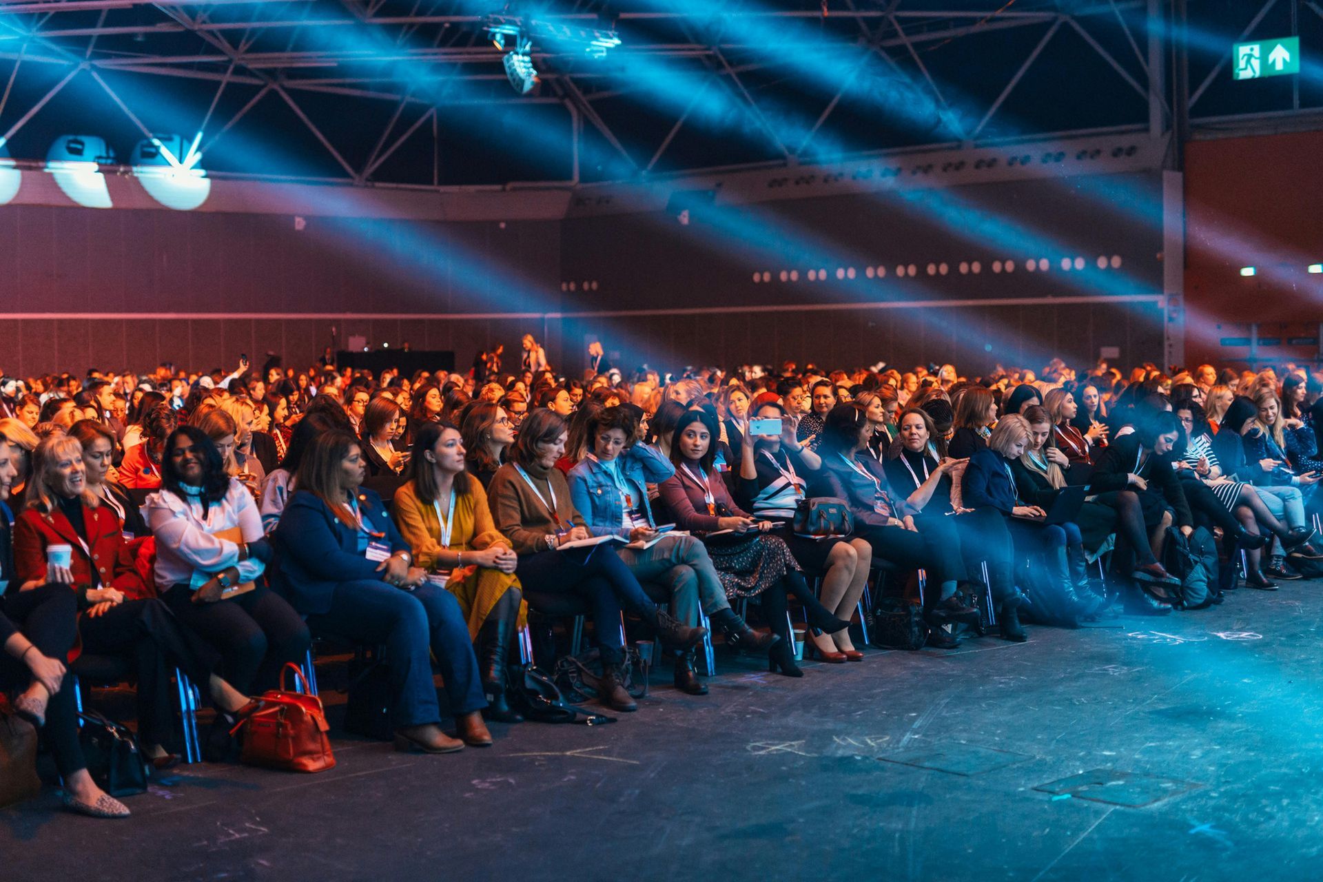 Publiek op een conferentie in een grote zaal, luisterend naar een presentatie. Podiumverlichting en schijnwerpers.