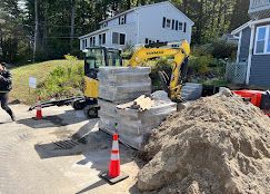 Construction site with yellow skid steer, stacked concrete blocks, and piles of dirt on a residential street
