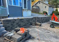 Construction site with stacked concrete retaining wall blocks beside a blue house and driveway equipment
