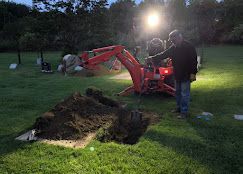 Person using a red backhoe at dusk to dig a hole in a grassy yard