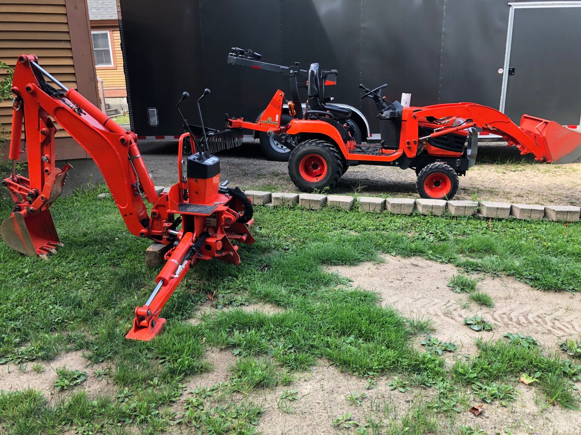 Two red mini excavators parked on grass near a building, one with a front loader bucket.