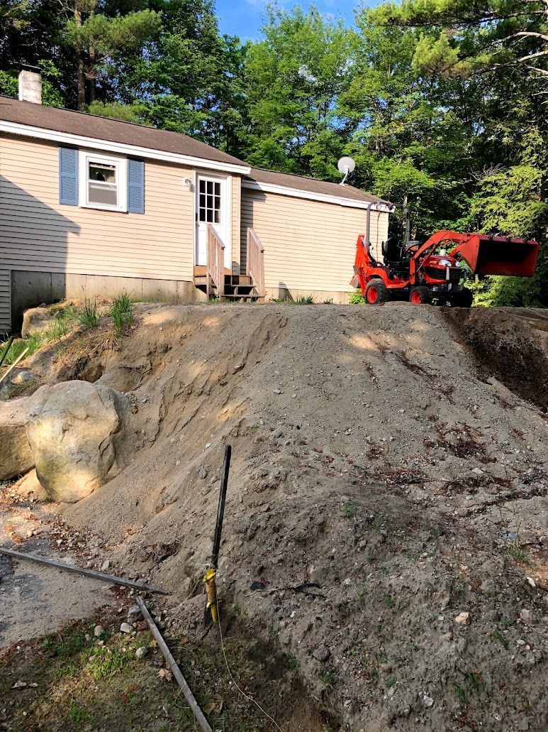 Dirt-covered yard with rocks, a beige house, and a red excavator on a sloped driveway under trees
