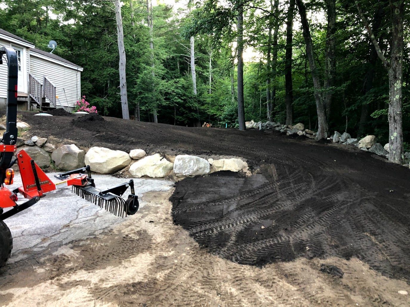 Freshly graded dirt driveway beside a house and trees, with construction equipment at the left edge.