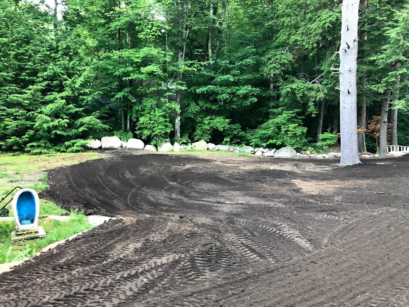Freshly graded dirt lot bordered by rocks and trees, with a blue plastic pet headstone on the left