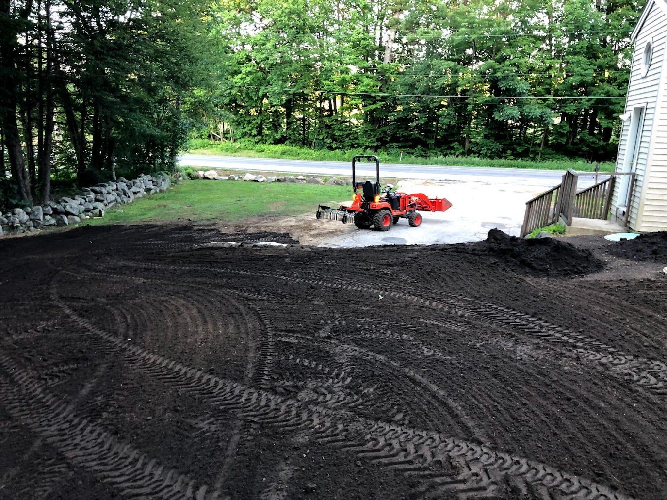 Small orange tractor leveling dark soil in a yard near a driveway and trees