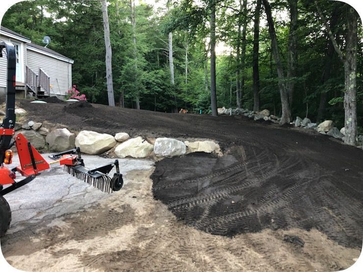 Freshly graded dirt driveway curving through a wooded yard beside a house and construction equipment
