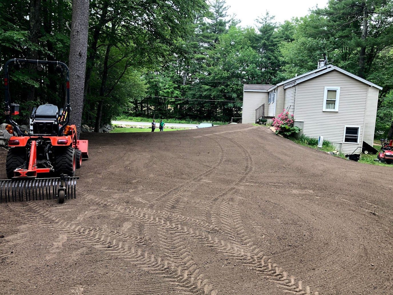 Freshly graded dirt driveway beside a white house, with a small tractor at left and trees in back