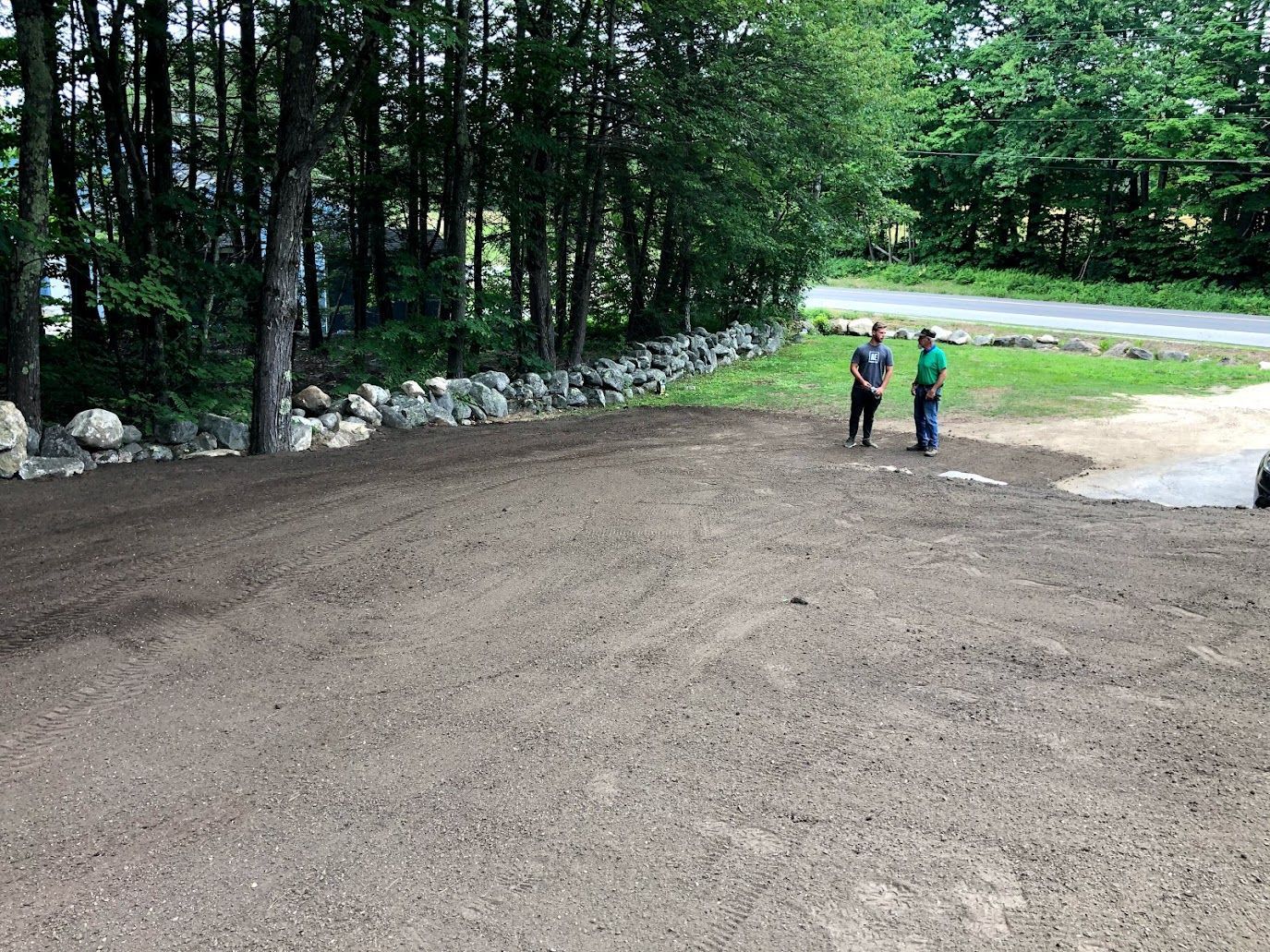 Two people standing on a freshly graded dirt area beside a wooded road