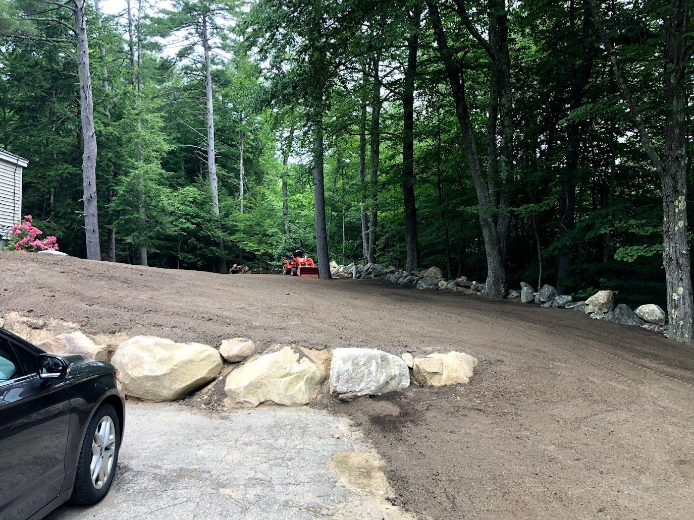 Gravel driveway beside a wooded yard with large rocks and a parked black car at left