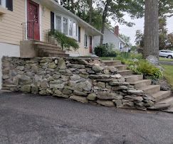 Stone retaining wall beside a house with front steps, along a sloped driveway.