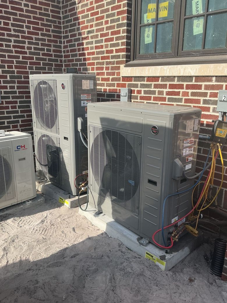 Two outdoor HVAC units beside a brick wall, with hoses and wiring attached.